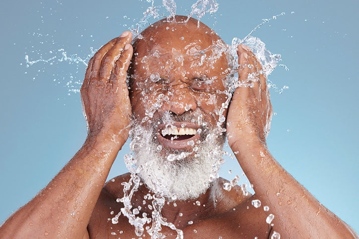 A close-up of a bald man with a white beard splashing water onto his face with both hands, eyes closed and teeth showing in an energized expression against a blue background