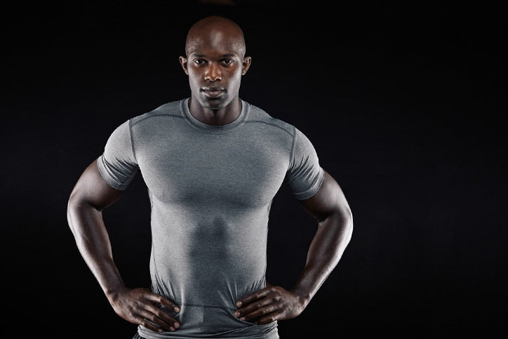 A muscular bald man in a tight grey athletic shirt standing with hands on his hips against a black background