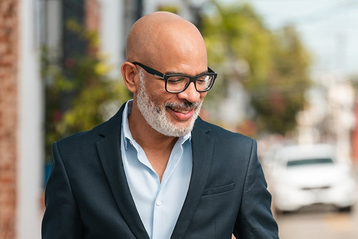 A smiling bald man with a white beard and glasses, wearing a navy blazer and light blue shirt, looking downward while walking outdoors.