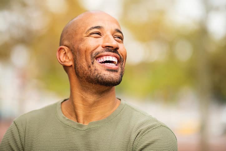 A laughing bald man with a beard wearing an olive green shirt, looking up and away against a blurred outdoor background