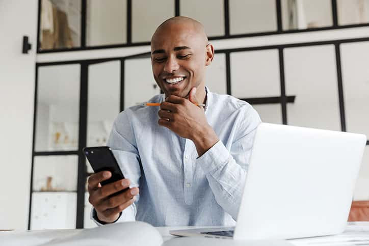 A smiling bald man sitting at a desk with a laptop, holding a smartphone in one hand and a pencil in the other
