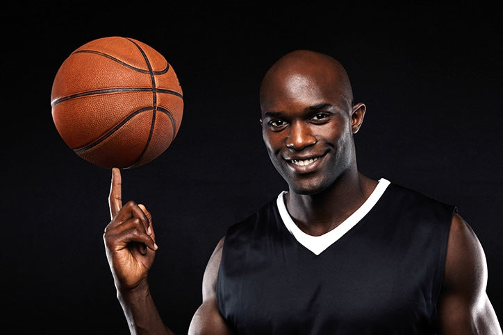 A smiling bald man in a black and white basketball jersey balancing a basketball on his fingertip against a solid black background