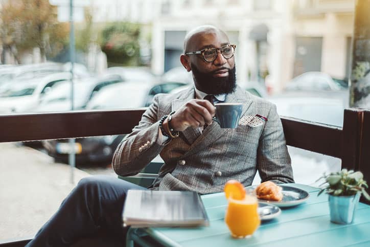 A bald African American man with a beard wearing a grey plaid suit and glasses, holding a coffee cup at an outdoor cafe table