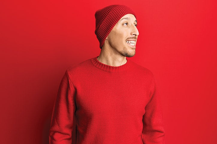 A smiling man wearing a red beanie and a red sweater, looking off to the side against a solid red background