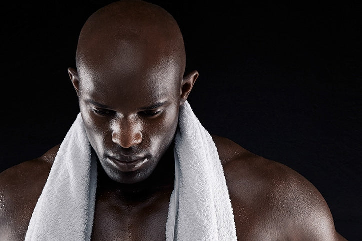 A bald African American man with damp, glistening skin and a white towel draped around his neck, looking downward against a black background