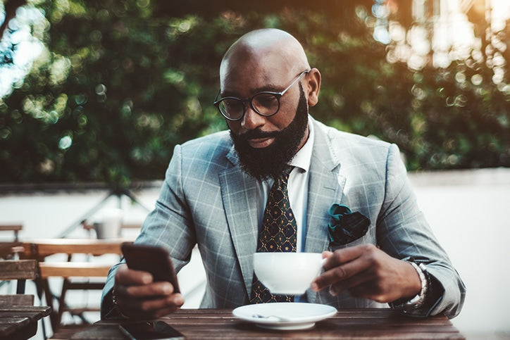 A bald African American man with a full beard and glasses, wearing a grey plaid suit and patterned tie, looking at a smartphone while holding a coffee cup at an outdoor table