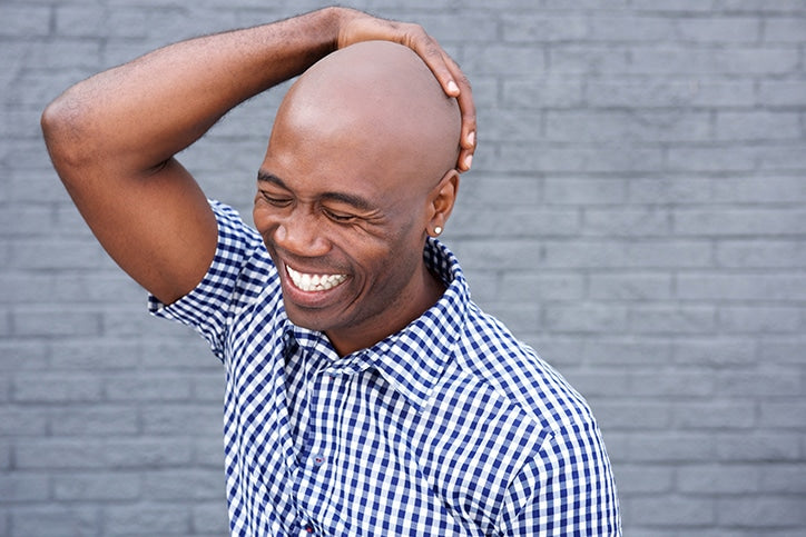A laughing bald African American man in a blue checkered shirt touching his smooth scalp against a gray brick wall
