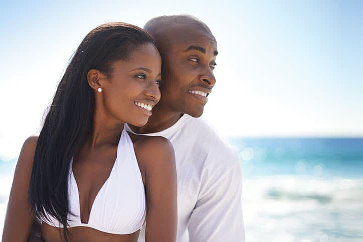 A smiling African American couple wearing white standing on a beach and looking out toward the ocean on a sunny day