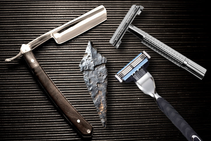 A collection of shaving tools including an ancient stone flint, a straight razor, a safety razor, and a modern multi-blade razor on a black textured background