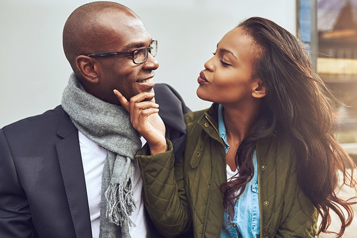 A African American woman leaning in to kiss a smiling bald man on the cheek while touching his chin outdoors