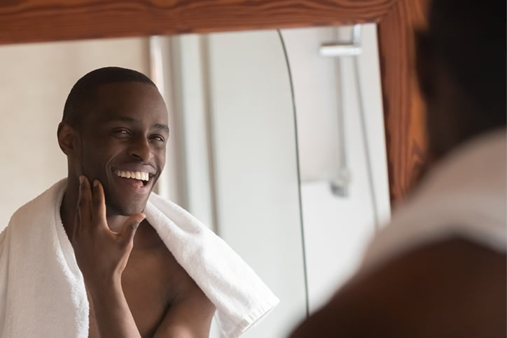 A smiling African American man with a towel around his shoulders looking in a mirror and touching his smooth, clean-shaven face