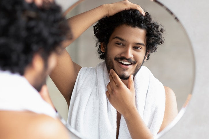 A smiling man with curly hair and a beard looking at his reflection in a round mirror while wearing a white towel around his neck and touching his chin
