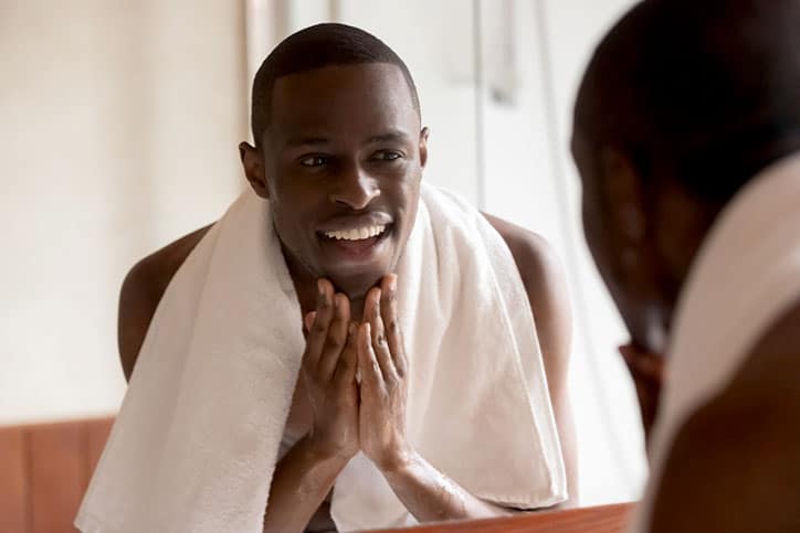A smiling man with a white towel around his shoulders looking at his reflection in a bathroom mirror while touching his clean-shaven jaw with both hands