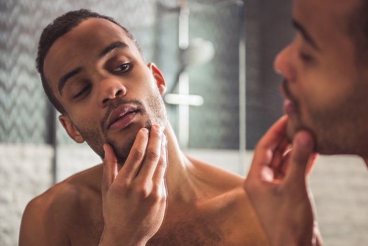 A man looking at his reflection in a bathroom mirror and touching the stubble on his chin