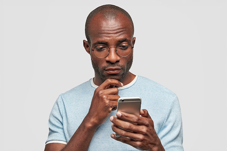 A man with glasses and a light blue t-shirt looking intently at a smartphone in his hands with a pensive expression, set against a plain white background