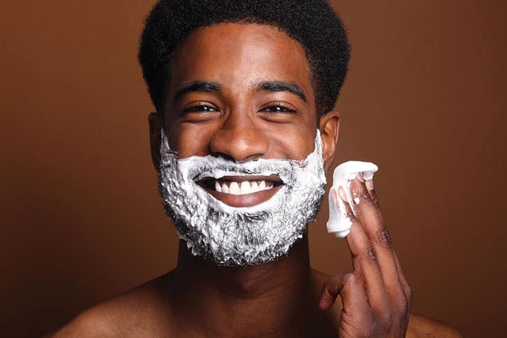A smiling African American man applying white shaving cream to his face and holding a dollop of foam on his fingertips against a brown background