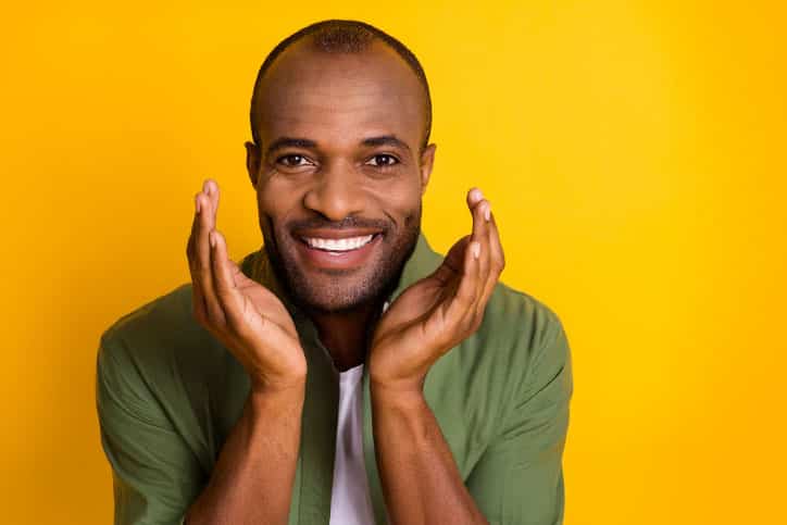 A smiling African American man in a green shirt framing his face with his hands against a bright yellow background