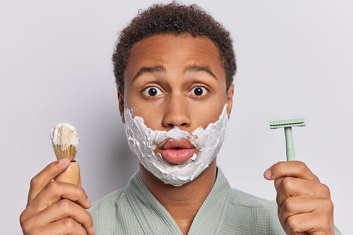 A young man with a surprised expression and shaving cream on his face, holding a shaving brush in one hand and a safety razor in the other against a light gray background