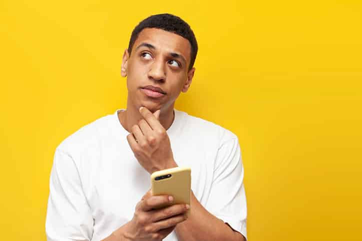 A man in a white t-shirt holding a yellow smartphone while looking up with a thoughtful expression against a bright yellow background