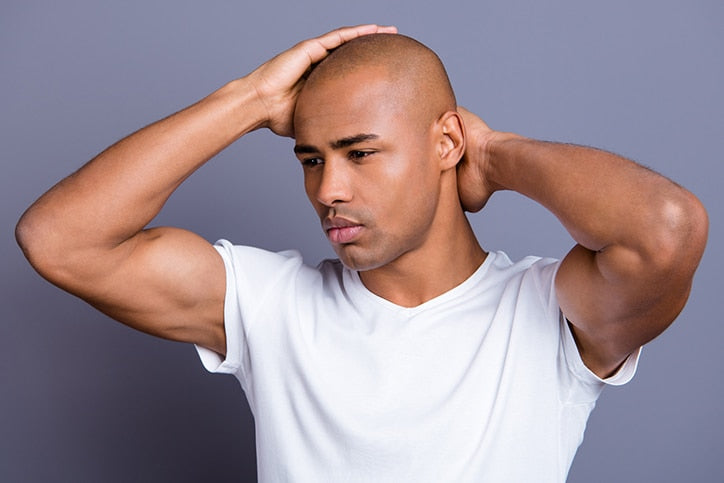 A man in a white t-shirt looking down thoughtfully while holding both hands behind his head and neck against a solid gray background
