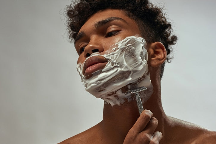 A man with curly hair using a safety razor to shave white shaving cream from his neck against a neutral background