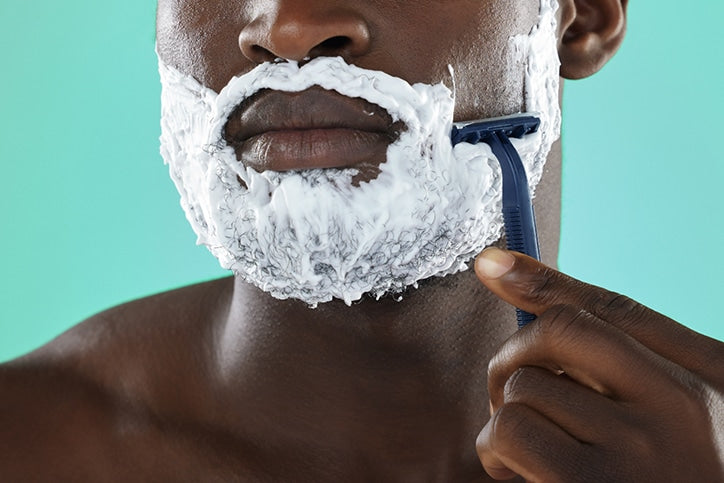 A close-up of a African American man using a blue disposable razor to shave white shaving cream from his cheek against a teal background