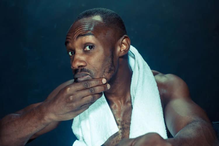 A man with a concerned expression holding his hand over his mouth and chin while looking into a mirror, with a white towel draped over his neck against a dark, moody background