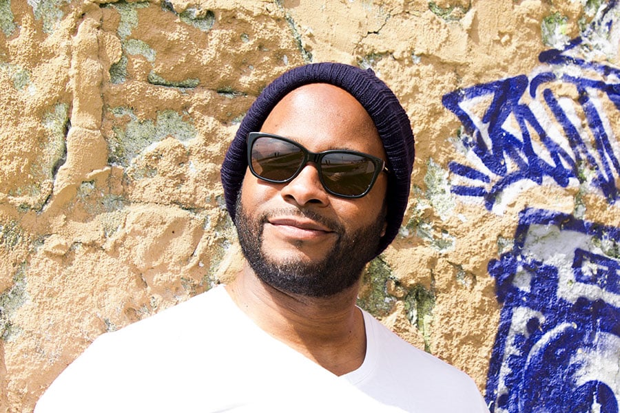 A man with a beard wearing a dark knit beanie and sunglasses, leaning against a textured wall with graffiti
