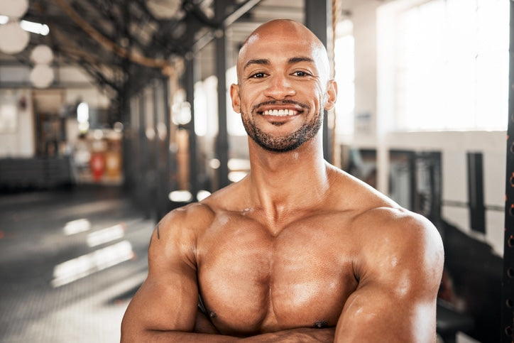 A smiling, muscular bald man posing with his arms crossed in a brightly lit gym with exercise equipment in the blurred background