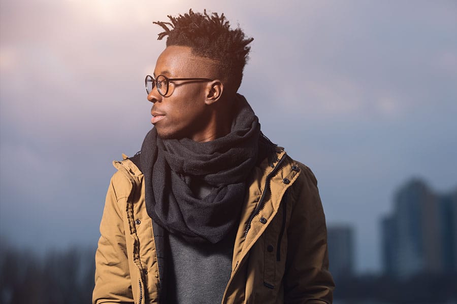 An African American man wearing a heavy winter jacket, a thick black scarf, and glasses, looking to the side against a cold, overcast sky