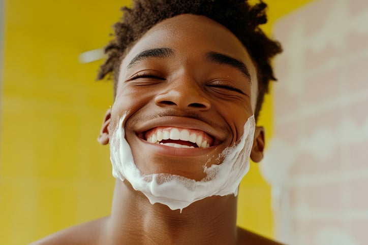 A close-up of a smiling young man with his eyes closed and white shaving cream covering his jawline and chin, set against a bright yellow background