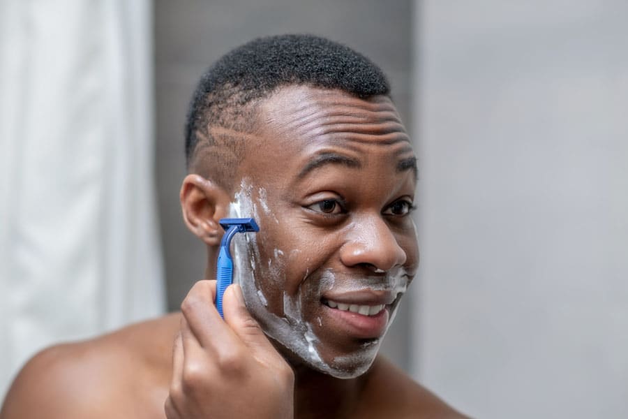 A man smiling as he uses a blue razor to shave his cheek, which is covered in white shaving gel, while looking into a mirror in a bathroom setting