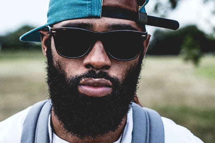 A close-up portrait of a man with a full, thick black beard wearing dark sunglasses and a teal baseball cap turned backward, set against a blurred outdoor park background