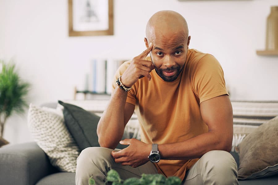 A bald African American man in a yellow t-shirt, sitting on a sofa and pointing to his smooth scalp