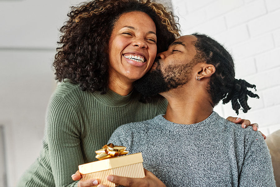A smiling African American woman receiving a gold gift box and a kiss on the cheek from a man with a beard