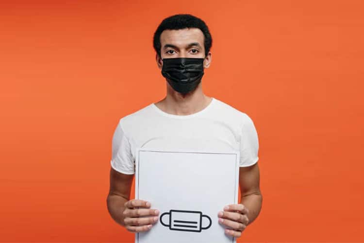 A man wearing a white t-shirt and a black face mask holding a white sign with a simple black icon of a face mask, set against a solid orange background