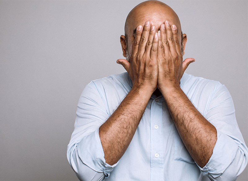 A man in a light blue button-down shirt covering his entire face with both hands against a grey background