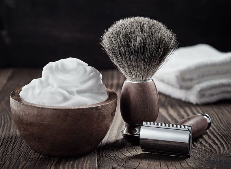 A traditional shaving kit on a dark wooden surface, featuring a wooden bowl overflowing with thick white shaving foam, a matching wooden-handled shaving brush, a safety razor, and a folded white towel in the background
