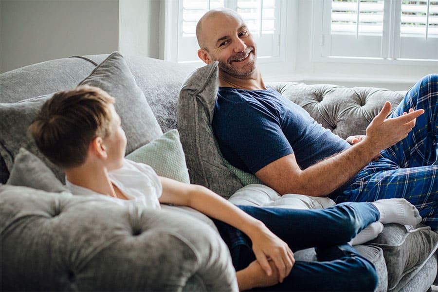 A stress-free bald father lying down on a couch, smiling while talking to his son.